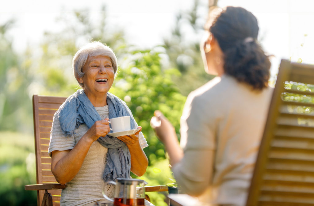 An older adult woman and her daughter sitting on lawn chairs outside, smiling and talking to each other while holding a warm beverage