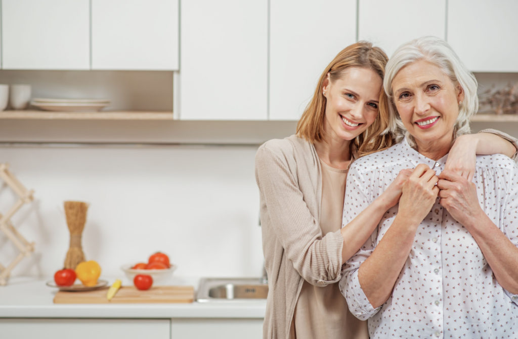 A young woman embracing her older adult mother in the kitchen, as they both look directly at the camera and smiling