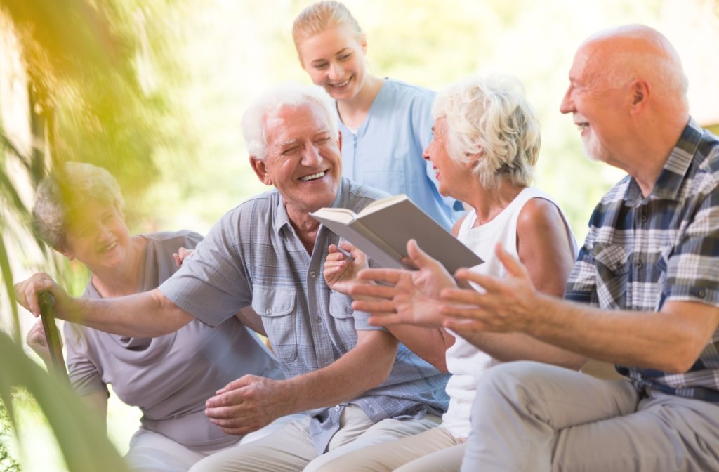 A senior man with a cane smiles and laughs while sitting outside with a nurse and other seniors.