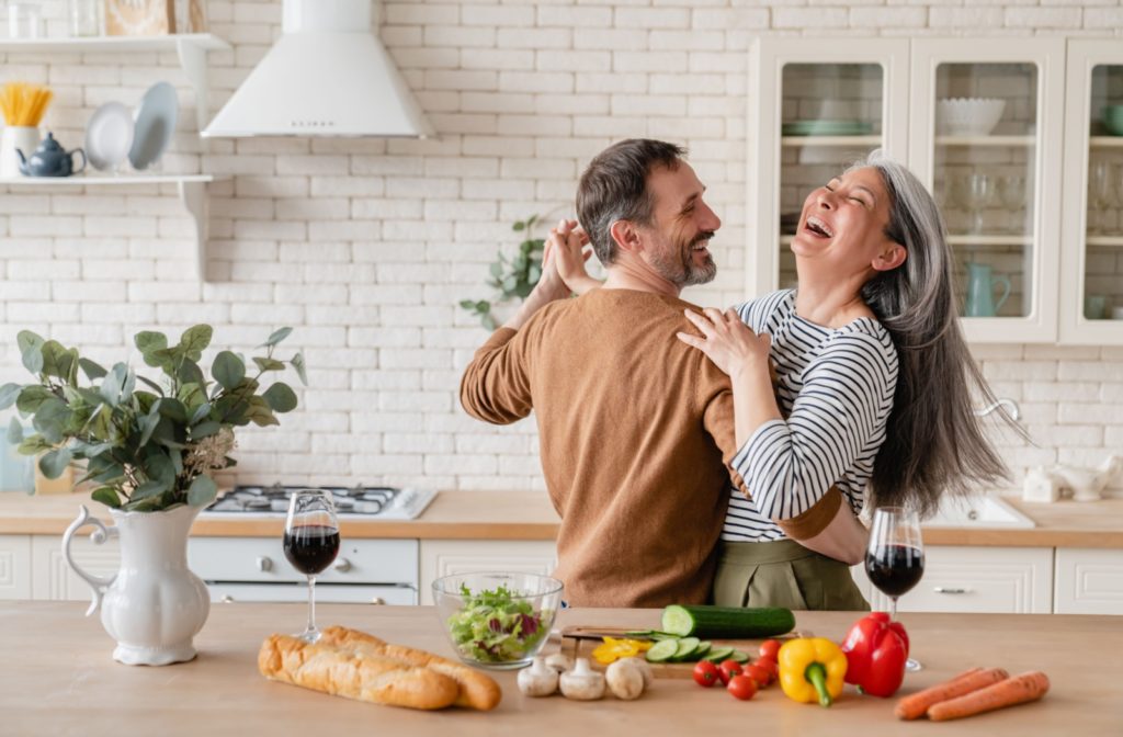 An older couple happily dancing in their kitchen surrounded by a variety of healthy foods.